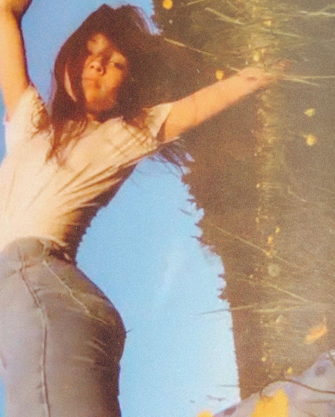 Young woman with long brown hair in a meadow against a blue sky, wearing a t-shirt and jeans, capturing a sense of carefree youth.