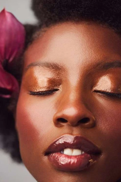 Close-up studio shot of a Black female model with glossy makeup, closed eyes, and a pink flower detail; beauty and cosmetic concept.