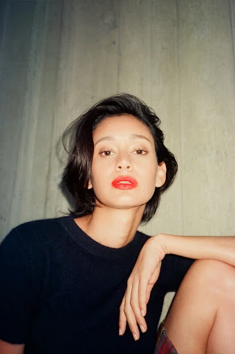 Portrait of a woman with dark short hair and bright red lipstick, gazing directly at the viewer in a natural-light indoor setting.