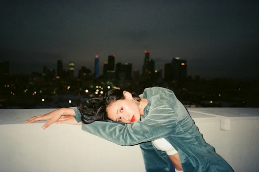 Woman in a green corduroy jacket resting her arms on a ledge with a distant city skyline at night