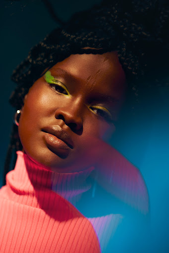 Portrait of a woman with green eyeshadow and pink turtleneck, shot in a photo studio with a blue background and soft lighting.