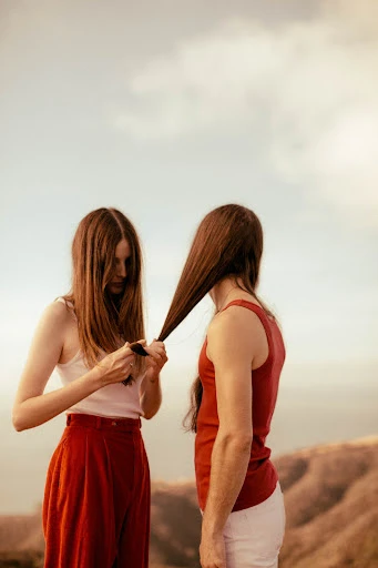 Two women with long brown hair in an outdoor setting. One woman is standing behind the other, holding her hair while standing on a hill.