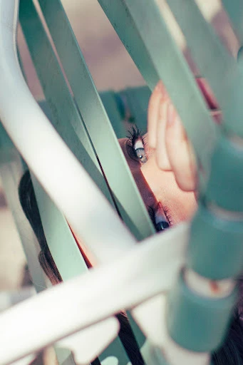 Close-up of a young adult female's face with one eye visible, partially obscured by blue and white metal bars in an outdoor setting.