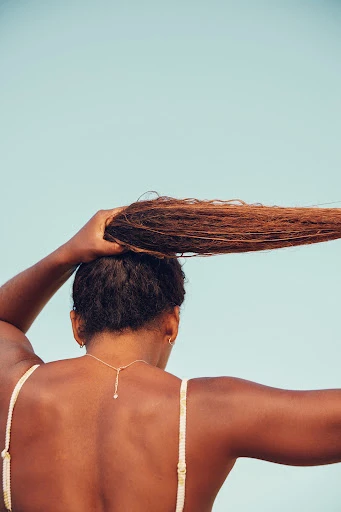 A dark-skinned woman with long brown hair raises it up against a light blue sky, showing her back and a gold necklace.