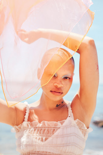 Portrait of a young adult woman with shaved head, wearing white top, holding sheer orange scarf, on a beach with ocean background on a sunny day.