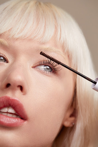 Close-up portrait of a blonde model applying brown mascara to her eyelashes in soft lighting