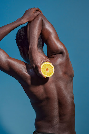 A dark-skinned man with a bare torso stretches with an orange half against a blue backdrop in a health and wellness theme.