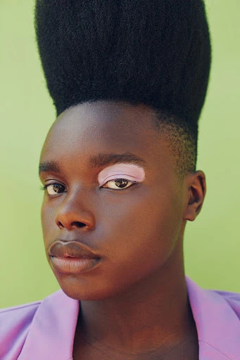 Close-up portrait of an adult with dark skin and a shaved head, wearing a purple blazer, with a light green background.