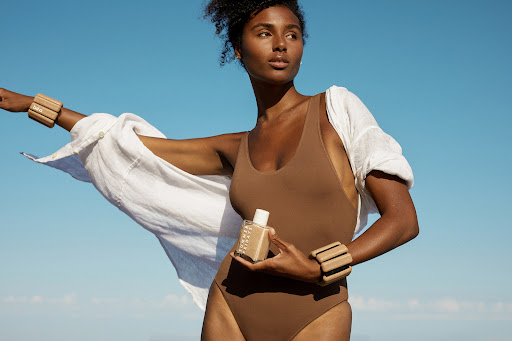 Attractive dark-haired woman in a brown swimsuit standing on a sunny beach holding product, wearing weights and white linen shirt.
