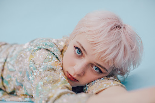 Close-up portrait of a young adult woman with blue eyes and pink hair lying down in a sequined jacket on a light blue background.