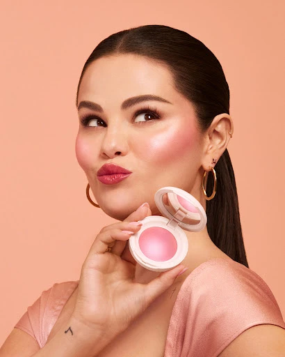 Close-up of a woman with fair skin holding a pink blush compact, wearing gold hoop earrings and a pink satin dress against peach backdrop.