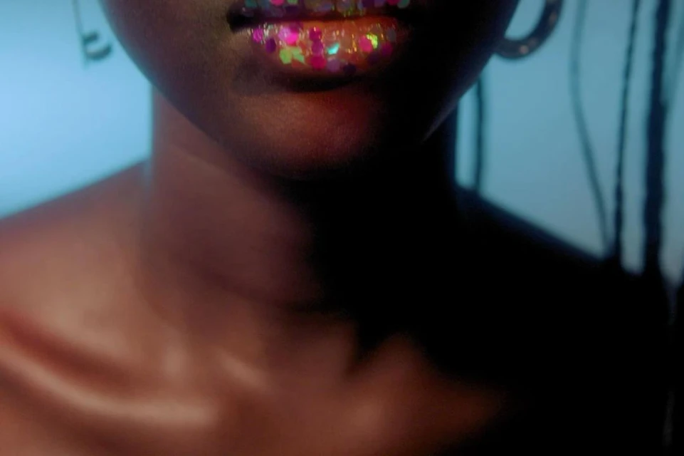 Close-up of an adult female with glitter on her lips, wearing hoop earrings against a blue background, with braids visible on the side.