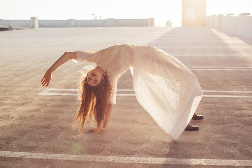 Young woman in flowing white dress strikes a backbend pose on a rooftop bathed in warm sunlight, displaying a blend of fashion and fitness.