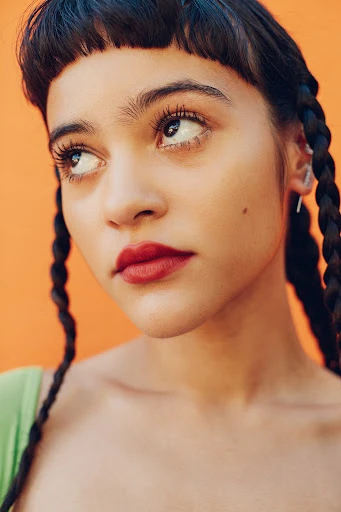 Portrait of a woman with braided hair, red lipstick, and fair skin against a plain orange backdrop, looking off-camera with a neutral expression.