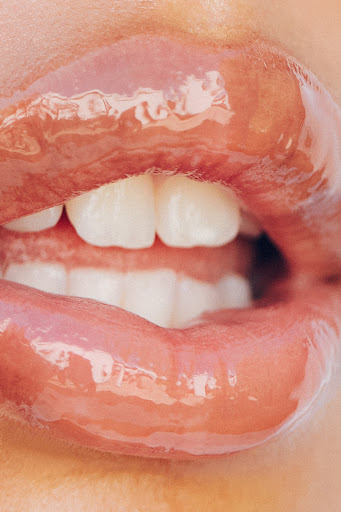 Close-up of an adult female's face, featuring glossy lips and white teeth, set against a blurred background. Beauty and fashion concept.