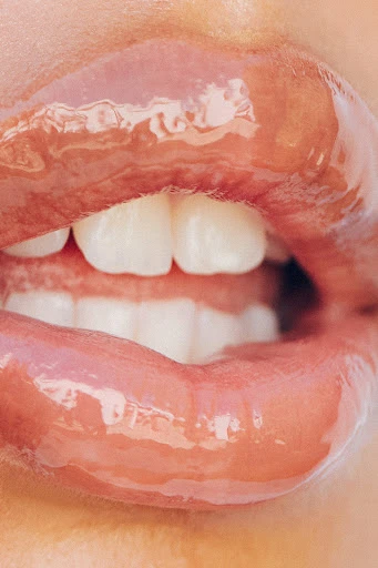 Close-up of an adult female's face, featuring glossy lips and white teeth, set against a blurred background. Beauty and fashion concept.