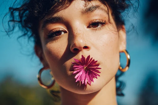 Close-up portrait of a woman holding a vibrant magenta flower between her lips against a clear blue sky