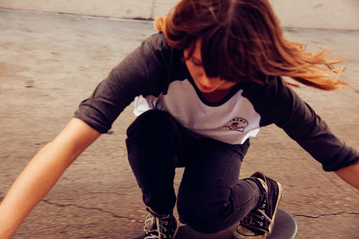 A youthful skateboarder with long brown hair in motion, captured in a dynamic medium shot on concrete in an outdoor setting, skateboarding.