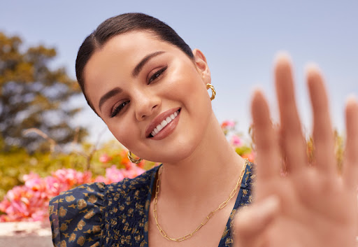 Close-up of Selena Gomez smiling outdoors, wearing a floral top and gold jewelry, with her hand raised towards the camera.