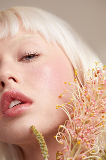 Close-up portrait of a fair-skinned adult female with blonde hair and pink lips, posed with a pink spider flower against a light beige background.