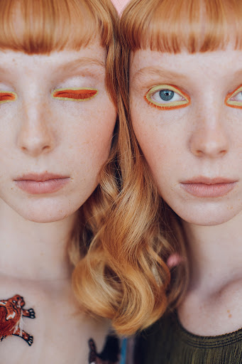 Close-up of two redhead women with striking yellow eyeliner, fair skin, and matching haircuts on a soft pink background.