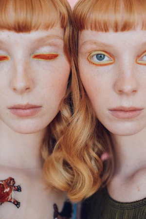Close-up of two redhead women with striking yellow eyeliner, fair skin, and matching haircuts on a soft pink background.
