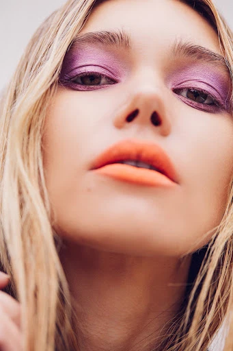 Close-up shot of a blonde woman with bright makeup featuring purple eyeshadow and coral lipstick in an eye-level portrait.