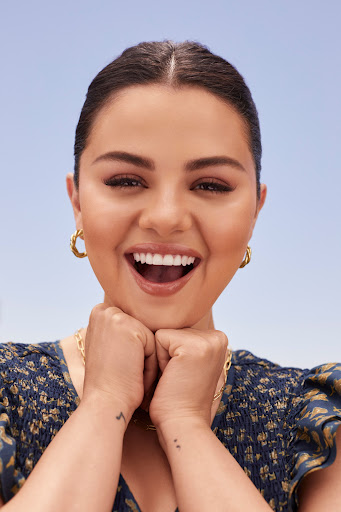 Close-up portrait of a smiling adult woman with brown hair and gold hoop earrings, wearing a dark-patterned dress against a blue sky.