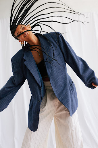 A smiling woman with box braids is wearing a pinstripe blazer and wide leg pants against a white backdrop in a fashion-forward pose.