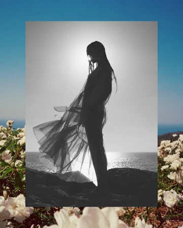 Monochrome image of a woman in flowing fabric, standing on a rocky outcrop with ocean and floral foreground, under a bright sky.