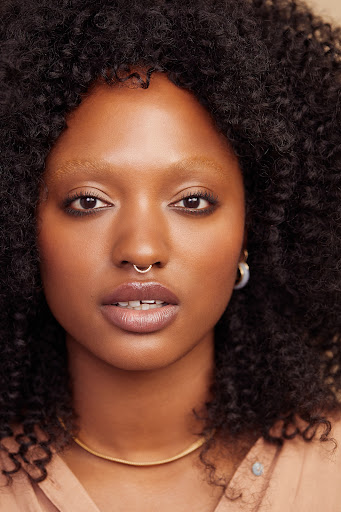 A portrait captures a Black woman in a blue top, smiling softly against a pink and orange background, highlighting her natural hair.