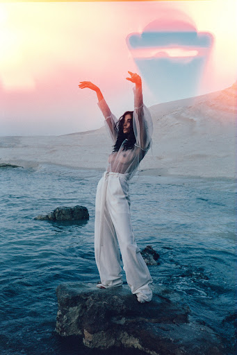 A woman in sheer top and white pants stands on a rock in a body of water with arms raised in front of a white mineral hill.