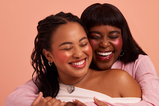 Two smiling women embrace in a close-up shot, showcasing makeup with a soft peach background. One woman wears a pink top.