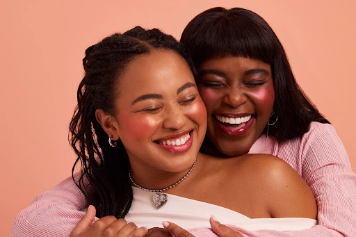 Two smiling women embrace in a close-up shot, showcasing makeup with a soft peach background. One woman wears a pink top.