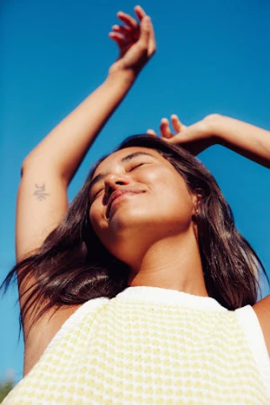 Woman with dark hair in a textured white top raising her arms against a clear blue sky by Kayla Varley