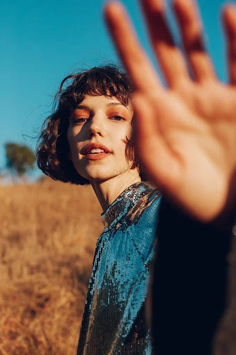 Portrait of a young adult female with short curly brown hair holding up her hand to the camera against a rural background.
