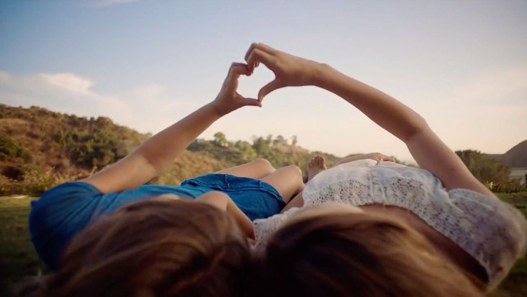 Two people lying on a grassy field making a heart shape with their hands, with a clear blue sky and distant trees in the background.