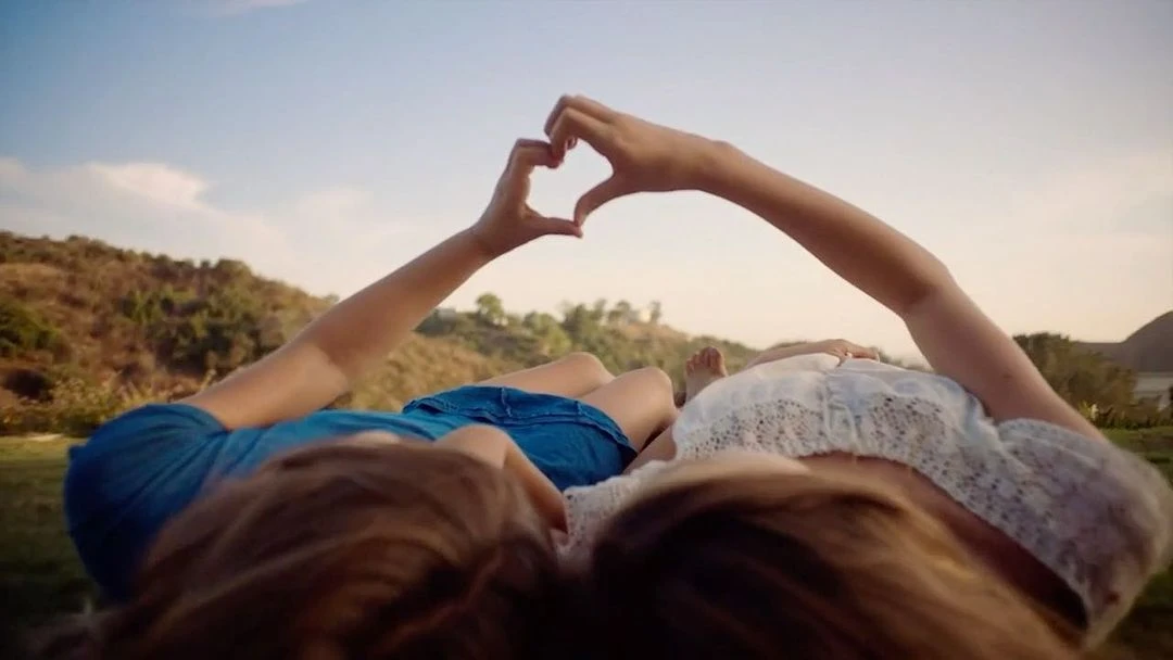 Two people lying on a grassy field making a heart shape with their hands, with a clear blue sky and distant trees in the background.