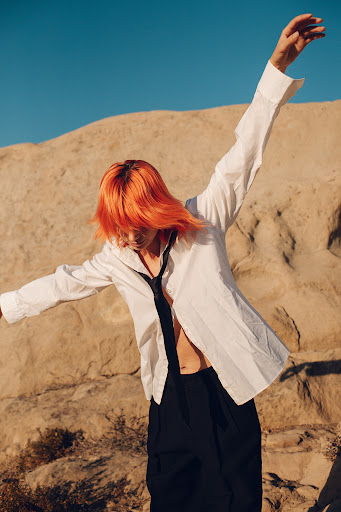 A person with vibrant orange hair in an unbuttoned white shirt and black pants stands in a desert with arms open under a clear blue sky.