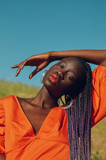 Adult Black female model with long braids poses with head tilted in a field against a blue sky. She wears an orange top.