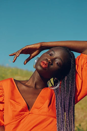 Adult Black female model with long braids poses with head tilted in a field against a blue sky. She wears an orange top.