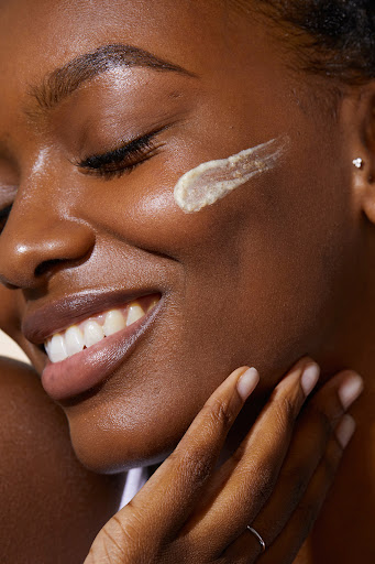 Close-up portrait of a smiling Black woman with skincare product swatched on her cheek by photographer Kayla Varley