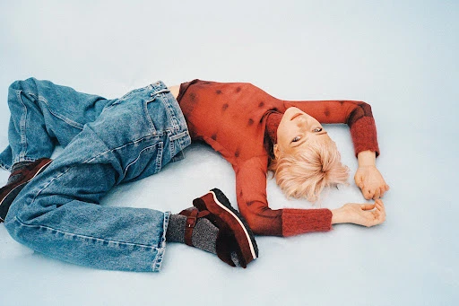Blonde-haired person in a red long-sleeved shirt and jeans poses while lying on a light-blue studio backdrop, facing the camera.