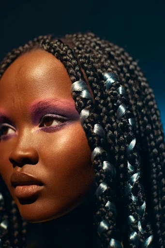 Portrait of an adult female with purple eyeshadow and a glossy lip wearing a braided hairstyle in front of a dark blue background.