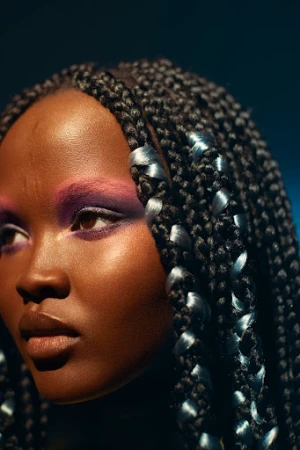 Portrait of an adult female with purple eyeshadow and a glossy lip wearing a braided hairstyle in front of a dark blue background.