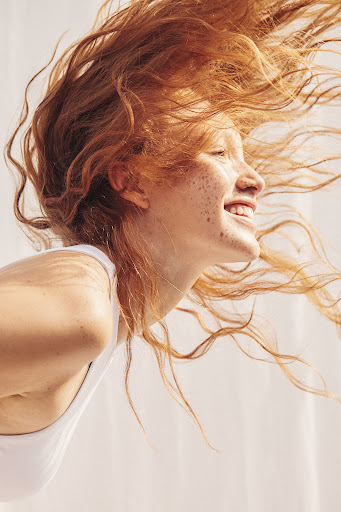 Close-up of a smiling adult female with freckles and red hair blowing in the wind, wearing a white top against a white background.