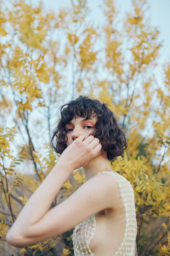 Portrait of a young woman with curly hair wearing a sequined top, standing in front of a tree with yellow leaves in natural light.