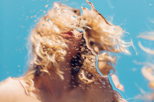 Close-up of an adult woman with blonde hair partially visible behind a textured surface with water droplets against a blue background.