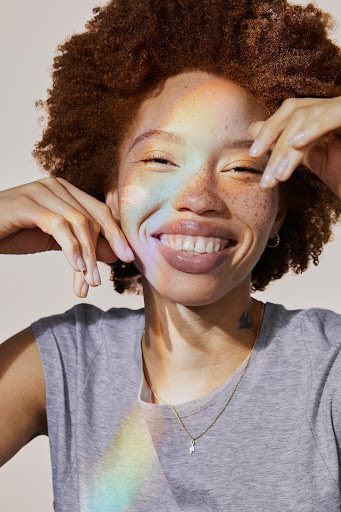 Smiling young woman with red hair and freckles poses indoors with light and shadow on her face and neck, wearing a gray top.
