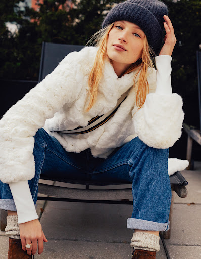 Blonde model poses in a white faux-fur jacket, jeans, and a gray beanie, seated outdoors on a black chair, in a medium portrait shot.